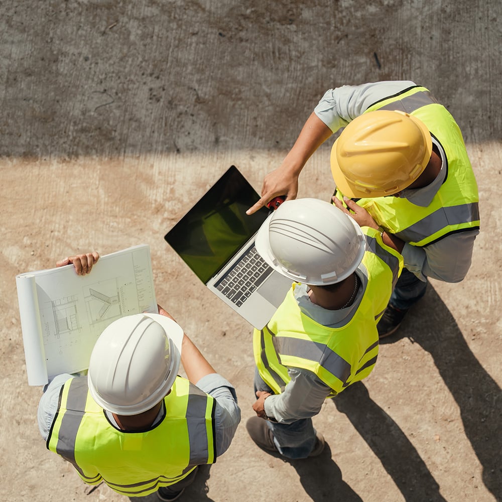 Three construction or project managers, wearing hard hats and high-visibility vests, reviewing plans on a laptop and blueprints at a construction site.